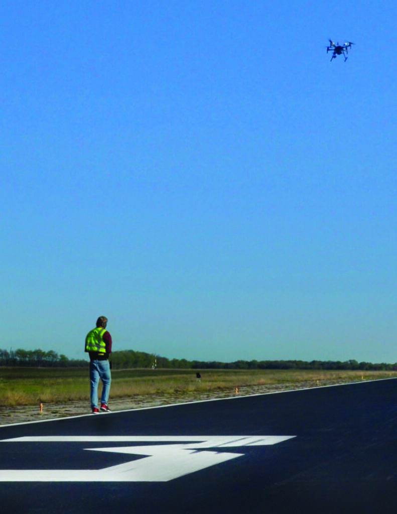 A person wearing a fluorescent safety vest is standing on the edge of an asphalt road, observing a drone flying in the clear blue sky.